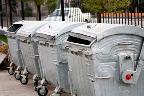 Four large metal rubbish bins with domed, grey, galvanized steel lids are aligned in a row on a paved surface, each equipped with black rubber wheels for mobility. The bins are positioned outdoors, with a slight tilt visible on the lid of the first bin from the right, revealing a dark opening beneath. The surface of the bins displays a textured, metallic finish with visible ridges and panel lines typical of industrial waste containers. In the background, there are blurred elements of a residential or commercial area, including trees with green foliage and a black metal fence, suggesting proximity to an urban environment in Southfields. The overall scene conveys a collection of waste receptacles prepared for rubbish disposal or collection, consistent with services like those offered by Garden Clearance Southfields for rubbish removal or garden clearance in the local area.