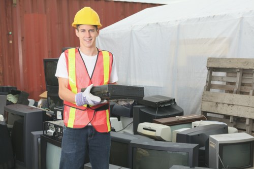 A young man wearing a yellow safety helmet, a high-visibility pink and yellow vest, and purple gloves is standing outdoors in front of a cluttered area with discarded electronic devices, including old computer monitors and printers, arranged on top of a wooden pallet and the ground. He is smiling and holding a black electronic device in both hands. Behind him, there is a white tent or cover, and a red wooden fence can be seen in the background. The scene suggests a rubbish removal or clearance activity, typical of garden clearance services in Southfields, where unwanted electronic waste is being prepared for collection or disposal.
