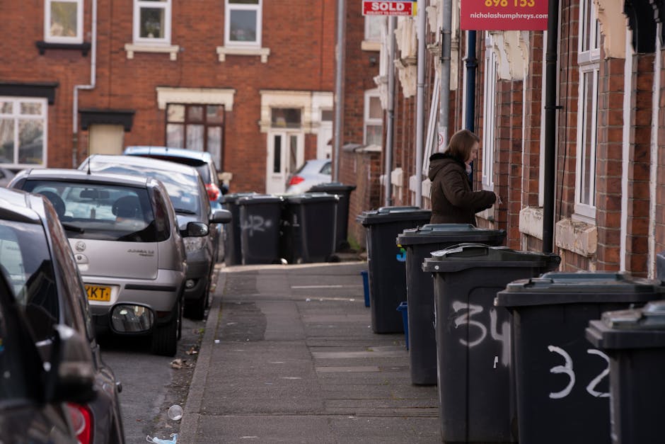 A person with short dark hair, wearing a black T-shirt with white text and light-colored gloves, is bent over a cylindrical metal rubbish bin on a paved sidewalk. The individual is holding a large whi